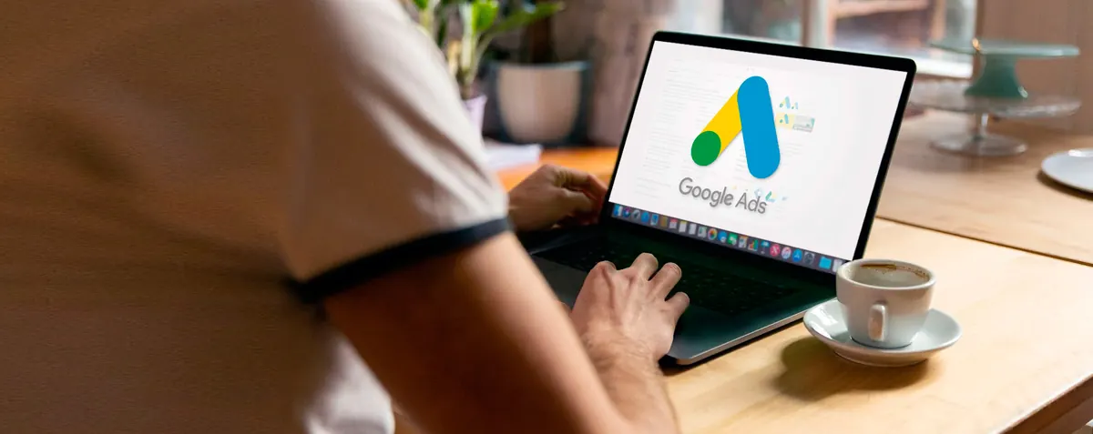 Persona trabaja en una portátil con el logotipo de Google Ads en pantalla, junto a una taza de café