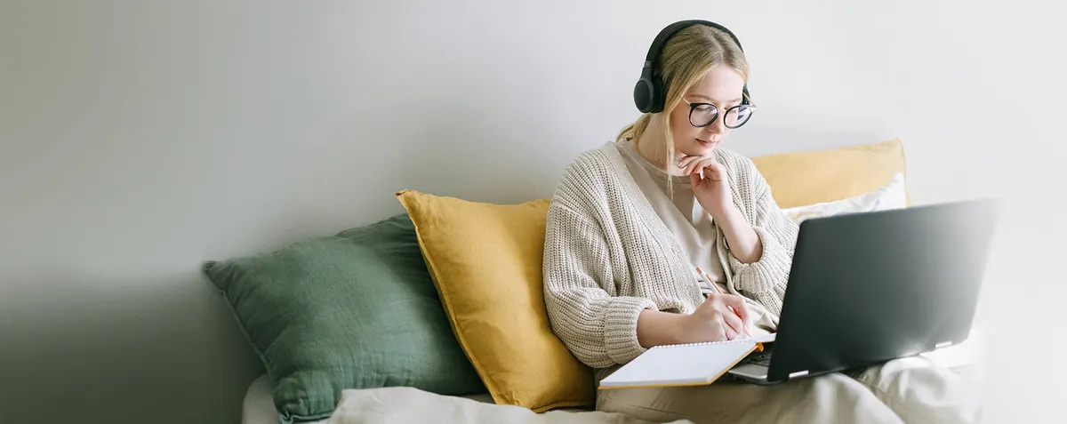 Mujer con audífonos y lentes toma notas junto a su portátil, sentada en un sofá con cojines